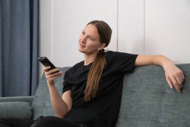 Genç bir kadın rahat bir kanepede uzanıp elinde uzaktan kumandayla şık oturma odasında televizyon seyrederken eğleniyor.