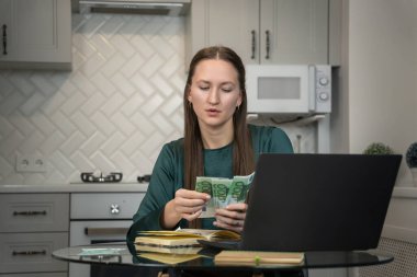 Ev bütçesini yöneten, dizüstü bilgisayar kullanırken avro banknotlarını sayan, mutfak masasında oturan ve mali planlama ve harcamalara odaklanan genç bir kadın.