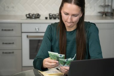 Genç kadın ev bütçesini hesaplıyor, modern mutfağındaki avro banknotlarını dikkatlice sayıyor, online bankacılık ve finansal yönetim için bir dizüstü bilgisayar kullanıyor.