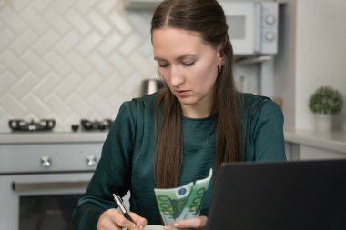 Avro banknotlarını elinde tutarken ve not defterine notlar yazarken mutfak masasında dizüstü bilgisayarla çalışırken ev bütçesini yöneten genç bir kadın.