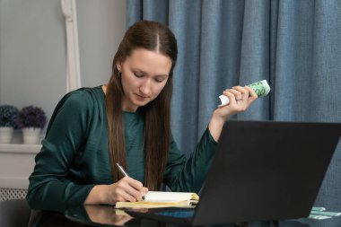 Genç bir kadın ev bütçesini yönetiyor, euro banknotlarını sayıyor ve modern bir mutfaktaki dizüstü bilgisayarla cam bir masada otururken deftere notlar yazıyor.