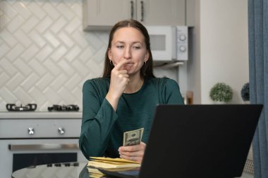 Genç bir kadın mutfak masasında oturmuş, dizüstü bilgisayarıyla uğraşırken nakit para sayıyor ve bir sonraki online alımını hesaplıyor, mali durumunu dengeliyor ve bütçesini planlıyordu.