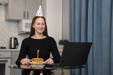 Parti şapkası takan genç bir kadın internetten doğum gününü kutluyor, dizüstü bilgisayarını kullanıyor, mutfakta pasta ve mumla oturuyor, arkadaşları ve ailesiyle video görüşmesi yapıyor.