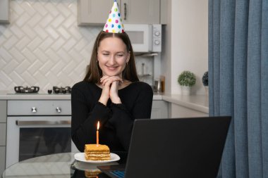 Parti şapkası takan genç bir kadın bilgisayarının önünde doğum günü pastası ve mumla doğum gününü internette kutluyor.