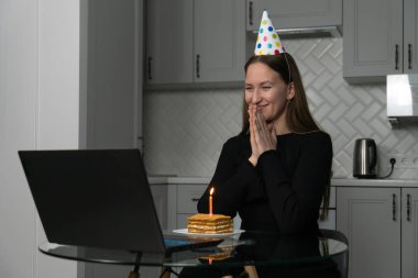 Parti şapkası takan genç bir kadın internette doğum gününü kutlayan dizüstü bilgisayara bakıp pasta ve mumla video görüşmesi yapıyor.