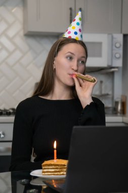 Doğum gününü internette kutlarken parti şapkası takan genç bir kadın. Mutfakta dizüstü bilgisayarla doğum günü pastası ve yanan mumla. Dikey fotoğraf
