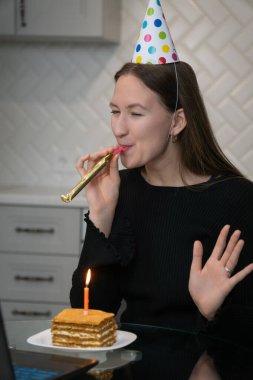 Doğum gününü internette kutlarken parti şapkası takan genç bir kadın. Mutfakta dizüstü bilgisayarla doğum günü pastası ve yanan mumla. Dikey fotoğraf