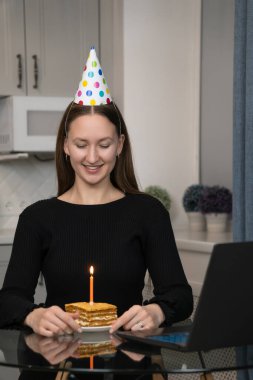 Doğum günü pastası ve mumla cam masada oturan parti şapkası takan genç bir kadın online doğum günü partisi kutlaması sırasında dilek tutuyor. Dikey fotoğraf