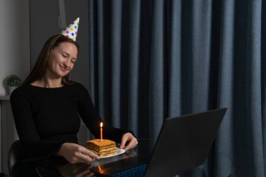 Doğum günü şapkası takan mutlu genç bir kadın pastaya mum üflemeden önce kapalı gözlerle bir dilek diliyor, evde tek başına kutlama yapıyor.
