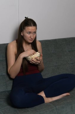 Uzun kahverengi saçlı genç bir kadın gri bir kanepede oturuyor, elinde cam bir kase patlamış mısır tutuyor ve hafif tiksindirici bir ifadeyle film izliyor. Dikey fotoğraf