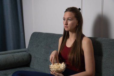 Oturma odasında sıkıcı bir film seyrederken cam bir kaseden patlamış mısır yiyen, rahat bir koltukta oturan genç bir kadın hayal kırıklığı ve ilgisizlik hissediyor.