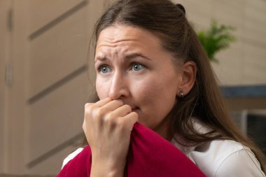 Stresli genç bir kadın kırmızı yastığı tutuyor ve gergin bir atmosferle çevrili parmaklarını endişeyle ısırıyor, işyerinde endişe ve duygusal gerginliği somutlaştırıyor.