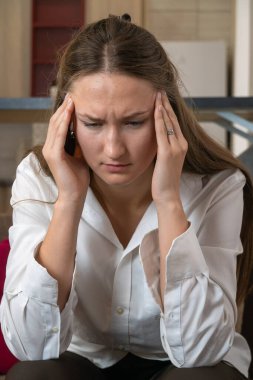 Beyaz gömlekli, migrenden muzdarip, baş ağrısı çeken, şakaklarına parmaklarıyla dokunan, evdeki bir kanepede oturan stresli genç bir kadın. Dikey fotoğraf