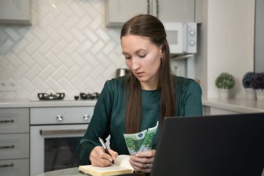 Kadın dikkatli bir şekilde ev ekonomisini yönetiyor. Titizlikle bir deftere harcamalarını yazıyor. Avro banknotlarını tutuyor ve mutfaktaki dizüstü bilgisayarını kullanıyor.