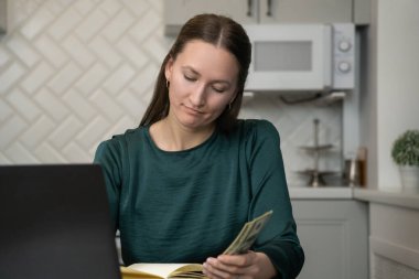 Bize dolar banknotları sayarak ev bütçesini yöneten, mutfaktaki cam masada oturan, dizüstü bilgisayarda çalışan ve not defterine notlar yazan genç bir kadın.
