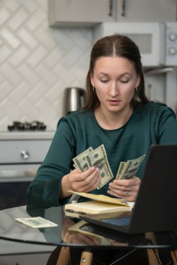 Ev ekonomisini yöneten genç bir kadın. Mutfak masasında dizüstü bilgisayar ve defterle oturup finansal sorumluluk ve ev bütçesini gösterirken bize dolar banknotlarını dikkatlice sayıyor.