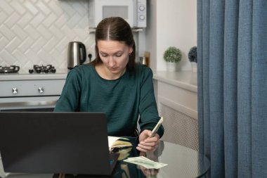 Genç bir kadın ev bütçesini yönetiyor, Amerikan dolarlarını sayıyor ve mutfak masasında oturmuş kişisel finans ve harcamalara odaklanırken dizüstü bilgisayarla çalışıyor.