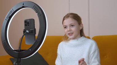 Genç bayan içerik yaratıcısı enerjik bir şekilde kameraya konuşuyor, profesyonel halka ışığıyla sarı koltukta oturuyor, ev stüdyosundaki dijital hikaye anlatımını somutlaştırıyor.