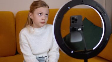 Beyaz kazaklı bir kız, tripodu ve halka ışığı olan akıllı bir telefondan çevrimiçi izleyicisi için ilgi çekici bir içerik oluşturan bir video kaydediyor.