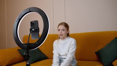 Genç nüfuzlu kişi, parlak sarı koltukta otururken video blogu kaydediyor. Profesyonel içerik yaratmak için akıllı telefon ve halka ışığını kullanıyor.