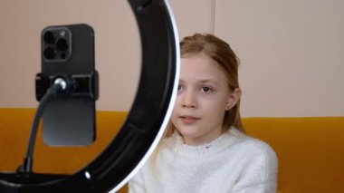 Beyaz kazaklı bir kız, tripodu ve halka ışığı olan akıllı bir telefondan çevrimiçi izleyicisi için ilgi çekici bir içerik oluşturan bir video kaydediyor.