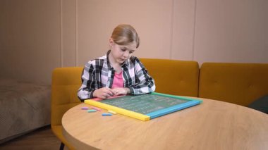 Genç bir ilkokul öğrencisi, çok renkli tebeşirle küçük bir tahtaya özenle sayı ve harf yazıyor ve aynı zamanda evde bilerek öğrenme sırasında konsantrasyonuyla konsantre oluyor.