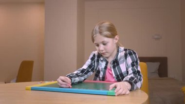 Odasındaki ahşap masada otururken yaratıcılığını ve konsantrasyonunu gösteren renkli manyetik tebeşirle manyetik tahtaya parlak çizgiler çizen liseli kız.
