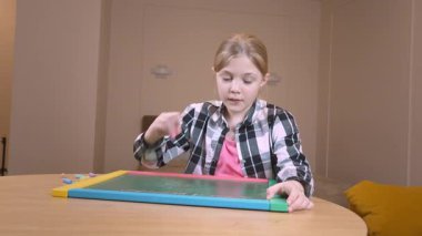 Odasındaki ahşap masada otururken yaratıcılığını ve konsantrasyonunu gösteren renkli manyetik tebeşirle manyetik tahtaya parlak çizgiler çizen liseli kız.