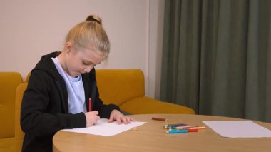 Sanatsal beceriler geliştiren odaklanmış bir öğrenci, rahat bir oturma odasında yuvarlak masada otururken renkli kalemleri kağıda dikkatlice çiziyor.