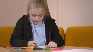 Sıcak oturma odasındaki yuvarlak ahşap masaya oturmuş renkli kalemlerle dikkatlice çizen kız çocukluk yaratıcılığını ve odaklanmış sanatsal ifadesini sergiliyor.