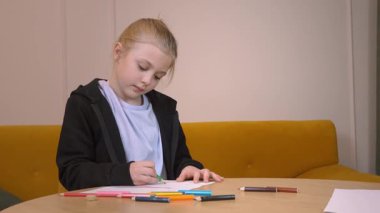 Sıcak oturma odasındaki yuvarlak ahşap masaya oturmuş renkli kalemlerle dikkatlice çizen kız çocukluk yaratıcılığını ve odaklanmış sanatsal ifadesini sergiliyor.