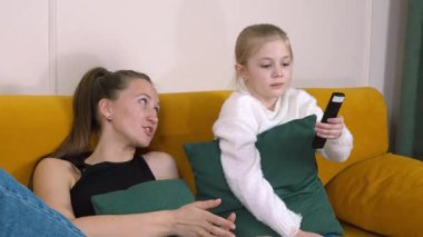 Anne ve kızı sarı bir kanepede oturmuş televizyon izliyorlar. Kız uzaktan kumandayı tutuyor ve anne ona bakarken tepkisini gösteriyor.