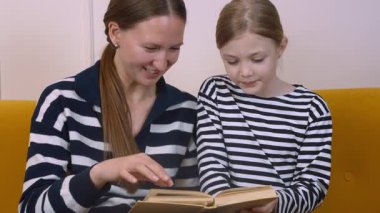 Annem, küçük kızına kitap okumasında yardım ediyor. Oturma odasındaki rahat sarı kanepede birlikte otururken kelimelere ve anlamlarına işaret ediyor.