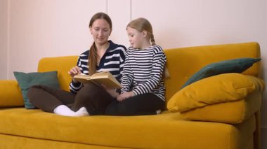 Annem, küçük kızına kitap okumasında yardım ediyor. Oturma odasındaki rahat sarı kanepede birlikte otururken kelimelere ve anlamlarına işaret ediyor.