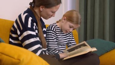 Annem, küçük kızına kitap okumasında yardım ediyor. Oturma odasındaki rahat sarı kanepede birlikte otururken kelimelere ve anlamlarına işaret ediyor.