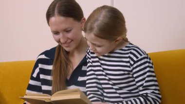 Annem, küçük kızına kitap okumasında yardım ediyor. Oturma odasındaki rahat sarı kanepede birlikte otururken kelimelere ve anlamlarına işaret ediyor.