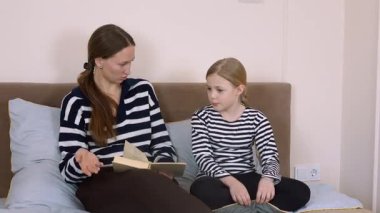 Anne kitap okuyor, kızıyla yatakta yan yana otururken hikâye detaylarını anlatıyor, sıcak yatak odasında öğrenme ve kaynaşma anını paylaşıyor.