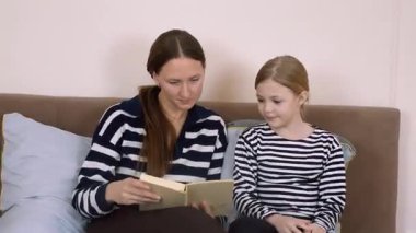 Anne kitap okuyor, kızıyla yatakta yan yana otururken hikâye detaylarını anlatıyor, sıcak yatak odasında öğrenme ve kaynaşma anını paylaşıyor.