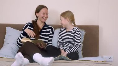 Anne kitap okuyor, kızıyla yatakta yan yana otururken hikâye detaylarını anlatıyor, sıcak yatak odasında öğrenme ve kaynaşma anını paylaşıyor.