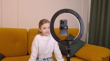 Hevesli genç kız çevrimiçi içerik yaratıyor, rahat bir koltukta otururken seyircilerle yakınlaşıyor, en uygun ışıklandırma için halka ışığını kullanıyor.