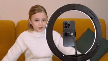 Beyaz kazaklı bir kız, tripodu ve halka ışığı olan akıllı bir telefondan çevrimiçi izleyicisi için ilgi çekici bir içerik oluşturan bir video kaydediyor.