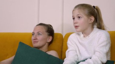 Anne sarı bir kanepede yatıyor ve kızı TV 'de korku filmi izliyor, yastıklara sarılıyor ve korku ve şaşkınlık duyguları gösteriyor.