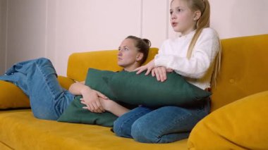 Anne sarı bir kanepede yatıyor ve kızı TV 'de korku filmi izliyor, yastıklara sarılıyor ve korku ve şaşkınlık duyguları gösteriyor.