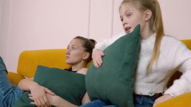 Anne sarı bir kanepede yatıyor ve kızı TV 'de korku filmi izliyor, yastıklara sarılıyor ve korku ve şaşkınlık duyguları gösteriyor.