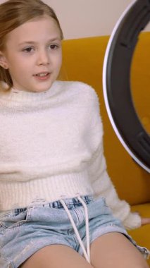 Genç kadın nüfuzlu kişi rahat bir koltukta oturuyor, profesyonel zil ışığıyla akıllı telefon kullanıyor ve modern oturma odasındaki sosyal medya kanalları için ilgi çekici video içeriği yaratıyor.