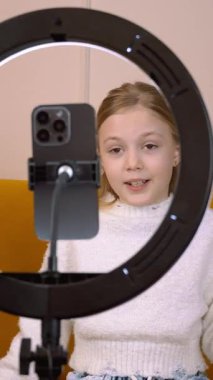 Genç kadın nüfuzlu kişi rahat bir koltukta oturuyor, profesyonel zil ışığıyla akıllı telefon kullanıyor ve modern oturma odasındaki sosyal medya kanalları için ilgi çekici video içeriği yaratıyor.