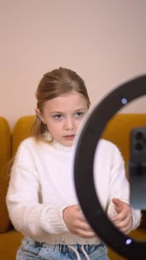 Genç kadın nüfuzlu kişi rahat bir koltukta oturuyor, profesyonel zil ışığıyla akıllı telefon kullanıyor ve modern oturma odasındaki sosyal medya kanalları için ilgi çekici video içeriği yaratıyor.