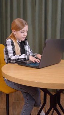 Odaklanmış kız öğrenci - Deochka, sarı kanepe ve modern ışıklandırma videosuyla rahat, aydınlık bir odada otururken dizüstü bilgisayarda yazmaya ve klavye üzerinde çalışmaya odaklandı.