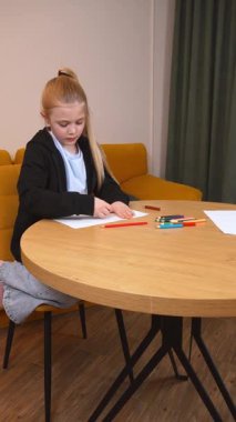 Yoğunlaşmış ilkokul öğrencisi yuvarlak bir masada renkli kalemlerle sarı kanepe ve yeşil perdeleri dikey olan rahat bir odada rahat bir şekilde oturan bir resim çiziyor.