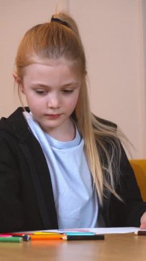 Renkli kalemle şirin çocuk çizimi. Evde resim çizen sevimli küçük bir anaokulu kızı. Küçük kız bir deftere bir şeyler yazıyor ve masa başında dikey bir videoda oturuyor.